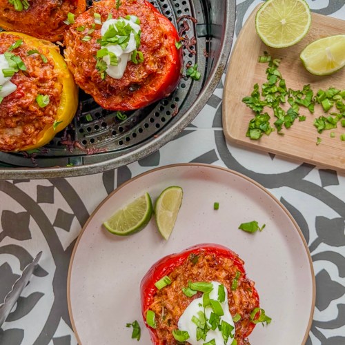 Air fryer ground chicken stuffed peppers in an air fryer basket with one on a plate and some fixings off to the side.