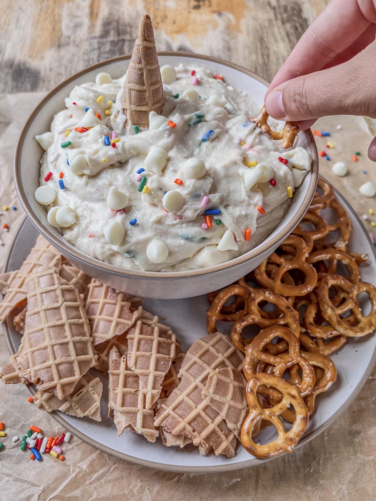 Cake batter cannoli dip with some rainbow sprinkles, waffle cone pieces, and pretzels scattered around