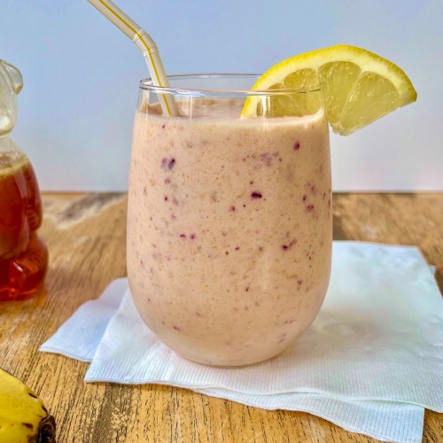 Cranberry clementine smoothie in a glass with a lemon wedge for garnish and some honey and fresh banana off to the side.