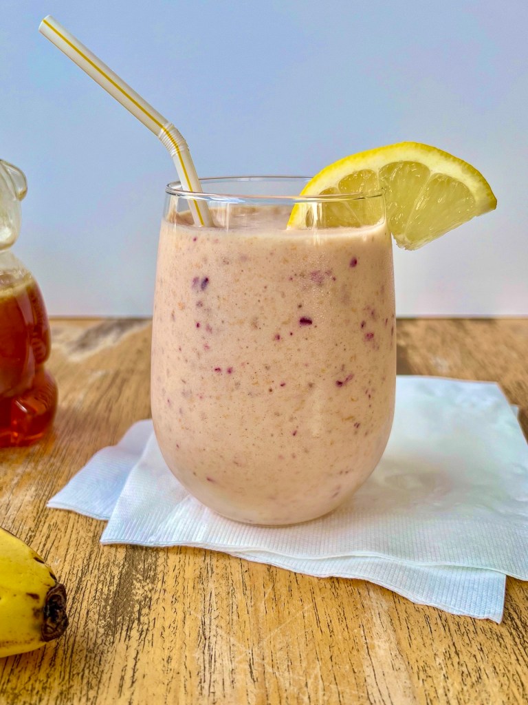 Cranberry clementine smoothie in a glass with a lemon wedge for garnish and some honey and fresh banana off to the side.