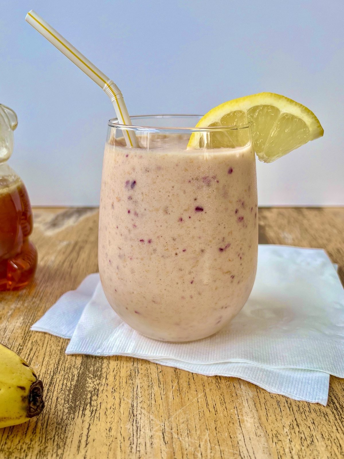 Cranberry clementine smoothie in a glass with a lemon wedge for garnish and some honey and fresh banana off to the side.