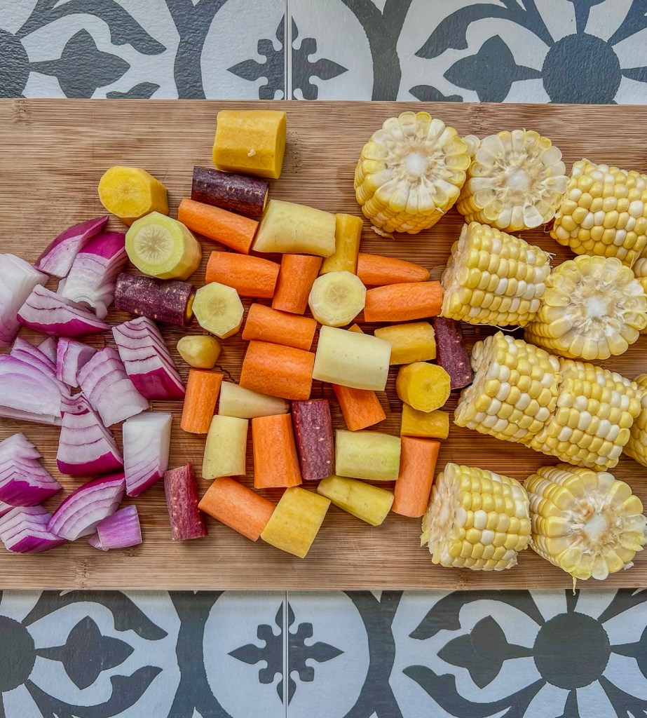 Red onion, rainbow carrots, and shucked corn on the cob prepped on a wooden cutting board.