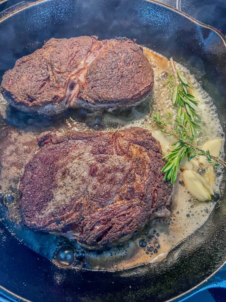 Steaks cooking in a cast iron with some newly added butter, crushed garlic cloves, fresh rosemary, and thyme.