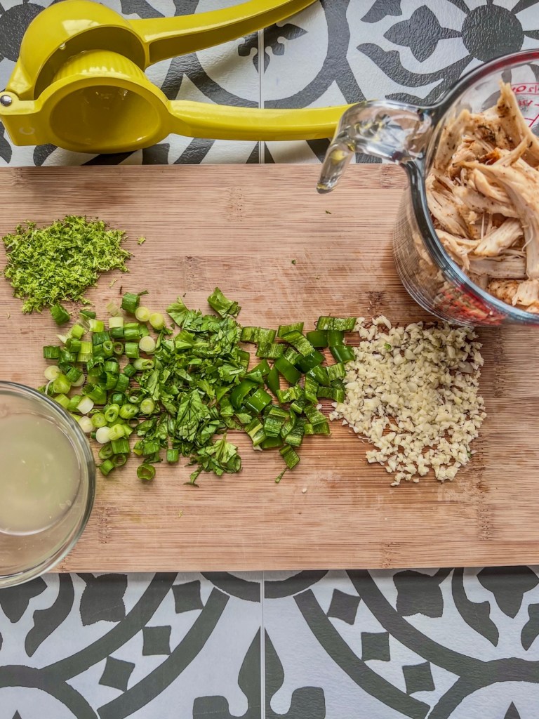 Green onion, cilantro, shredded chicken, garlic, lime juice and zest prepped on a cutting board.