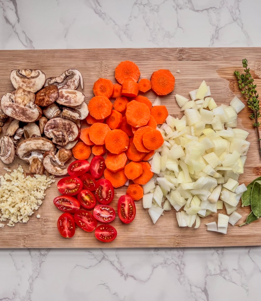 Onion, carrots, mushrooms, garlic, thyme, and tomato prepped on a wooden cutting board.