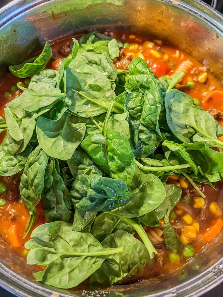 Ground beef veggie soup in a pot with some newly added spinach.