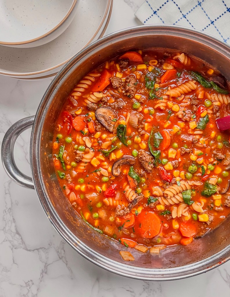 Rotini and beef vegetable soup fresh off the stove with some bowls stacked up nearby.