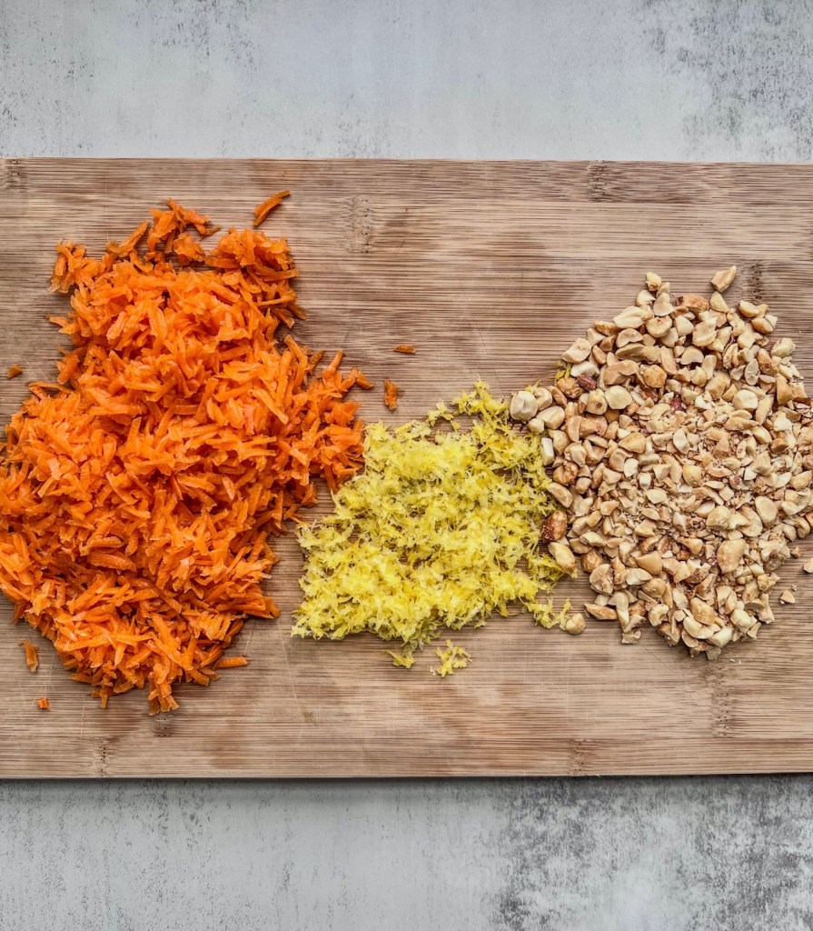 Shredded carrots, zested lemon, and chopped peanuts on a wooden cutting board.