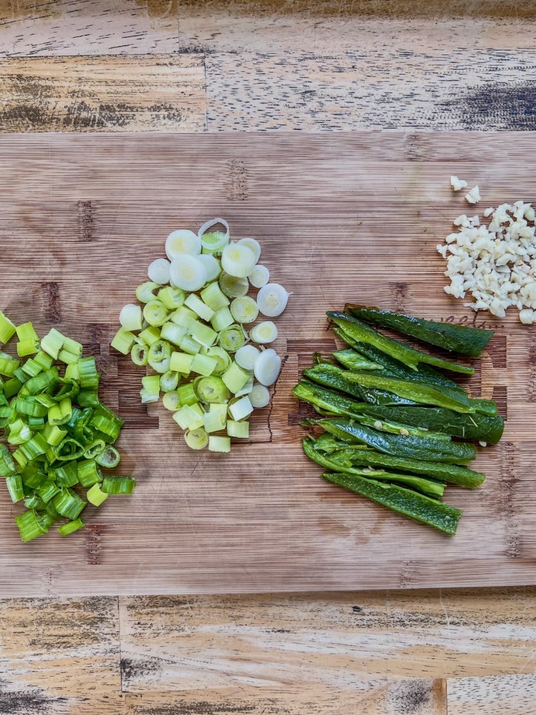 Green onions, broiled and sliced jalapeños, and garlic prepped on a cutting board.