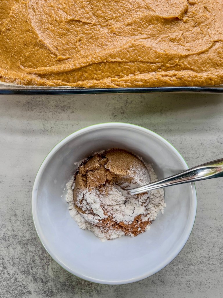 A spiced crumble topping being made in a small bowl with a large baking pan filled with pumpkin cake batter behind.