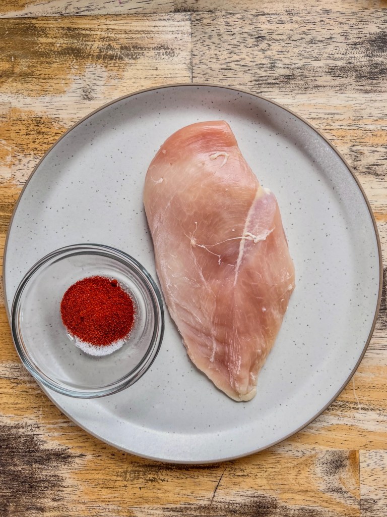 A chicken breast on a plate with a small dish full of spices next to it.