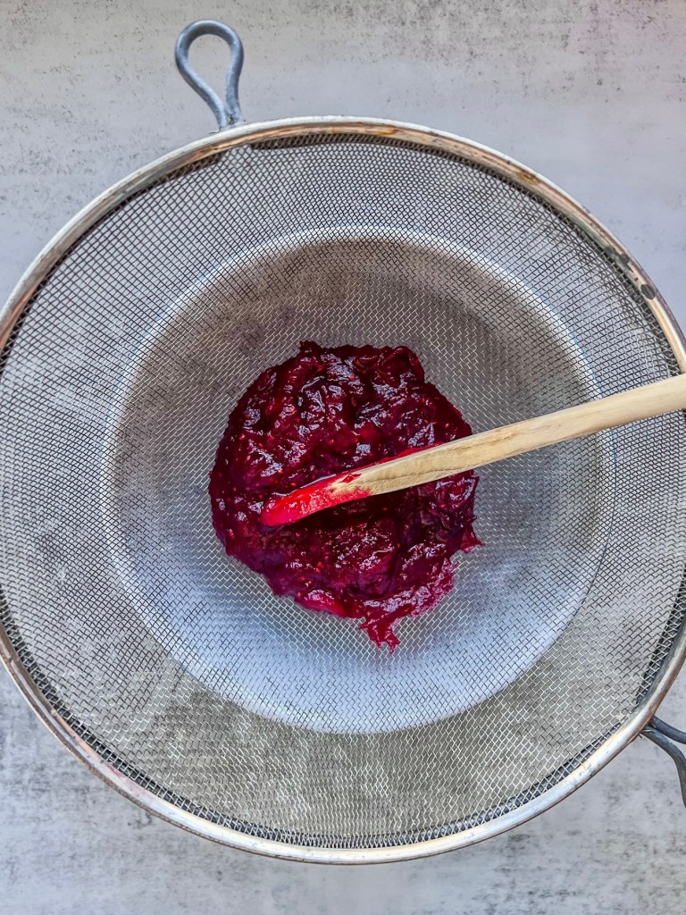 Cranberry compote being strained through a fine mesh sieve into a mixing bowl.