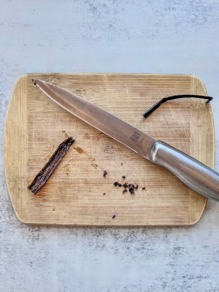 A vanilla bean that's been cut in half and had the seeds scraped out on a wooden cutting board.