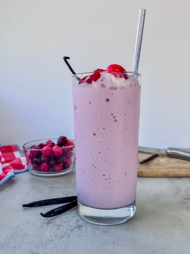 A cranberry vanilla bean milkshake in a large glass with some whipped cream, cranberry reduction, fresh cranberries, and a vanilla bean on top.