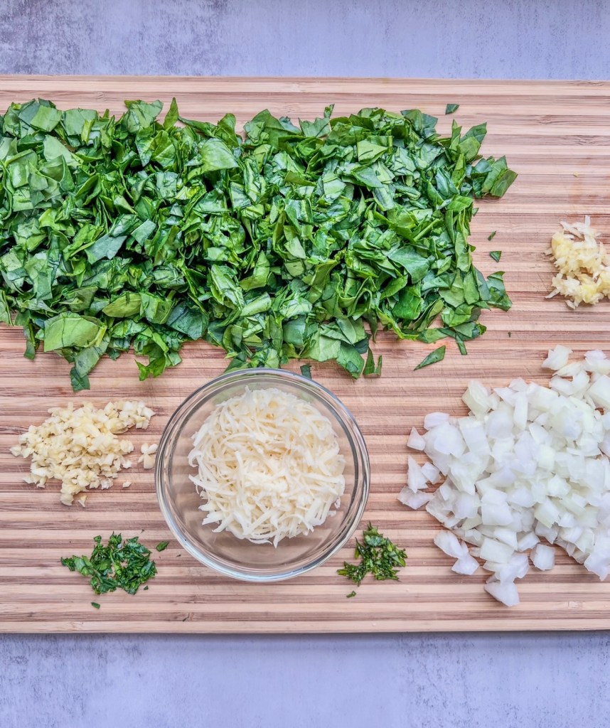Spinach, onion, garlic, basil, and fresh cheese prepped on a cutting board.