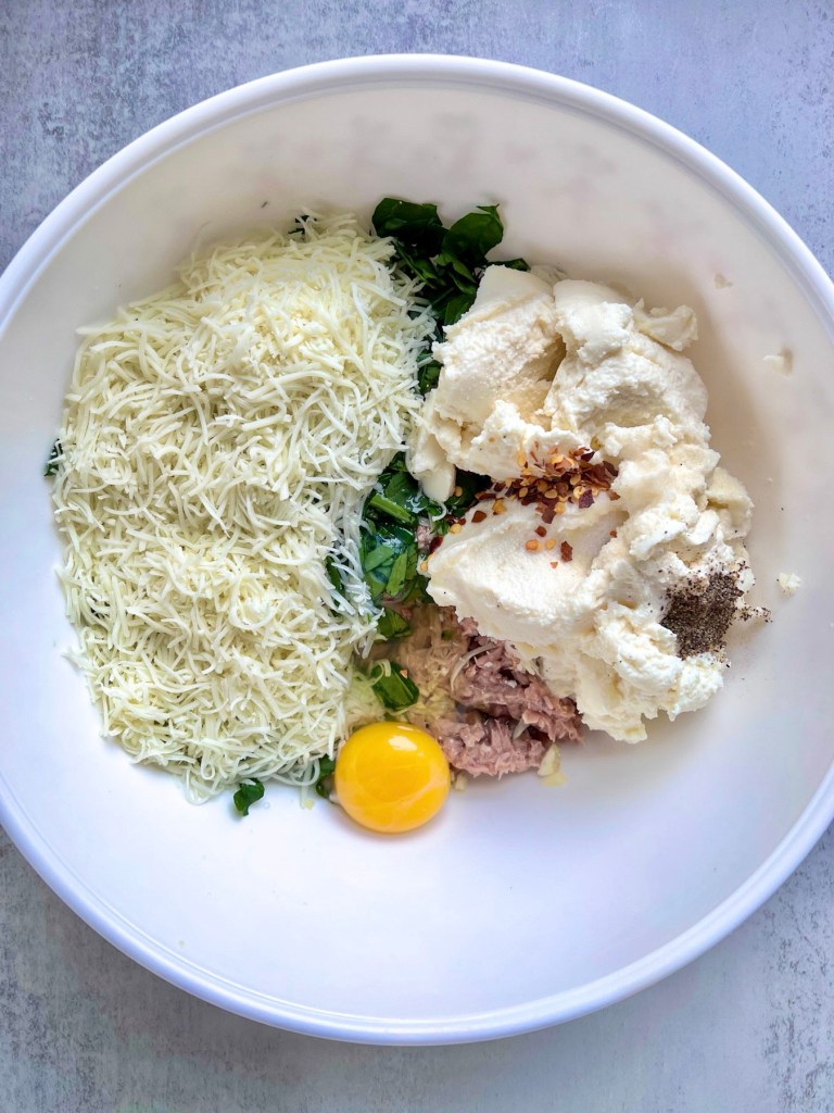 A bowl filled with tuna, garlic, cheese, spices, egg, and freshly chopped spinach.