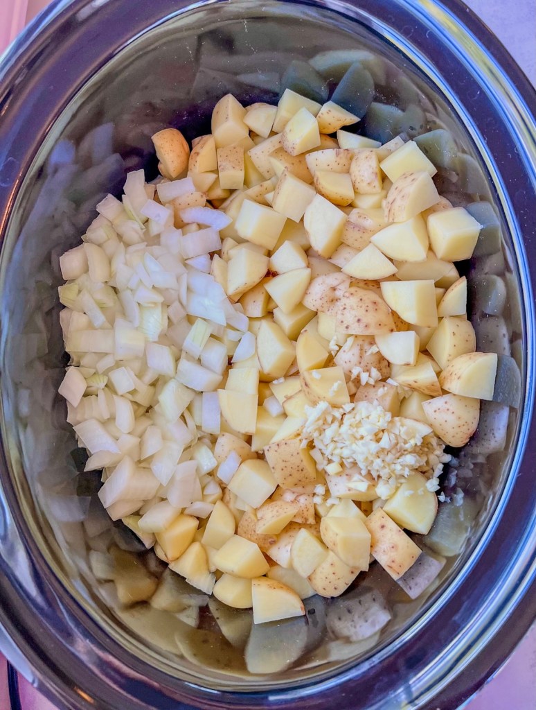Potatoes in a large crockpot with some onion and garlic scattered on top.